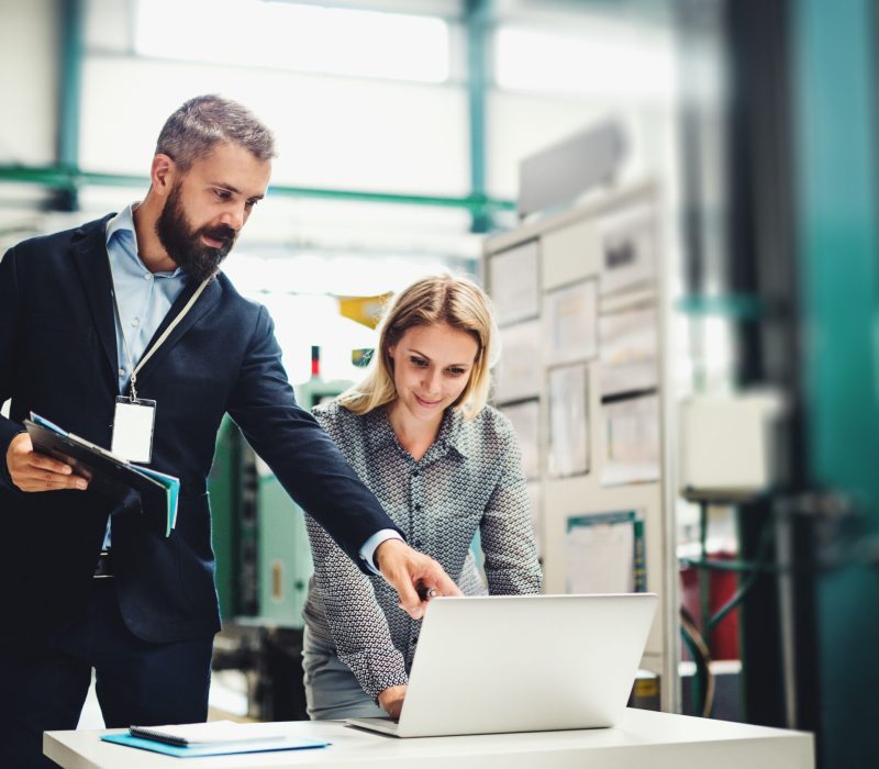 A portrait of a serious mature industrial man and woman engineer with laptop in a factory, working.